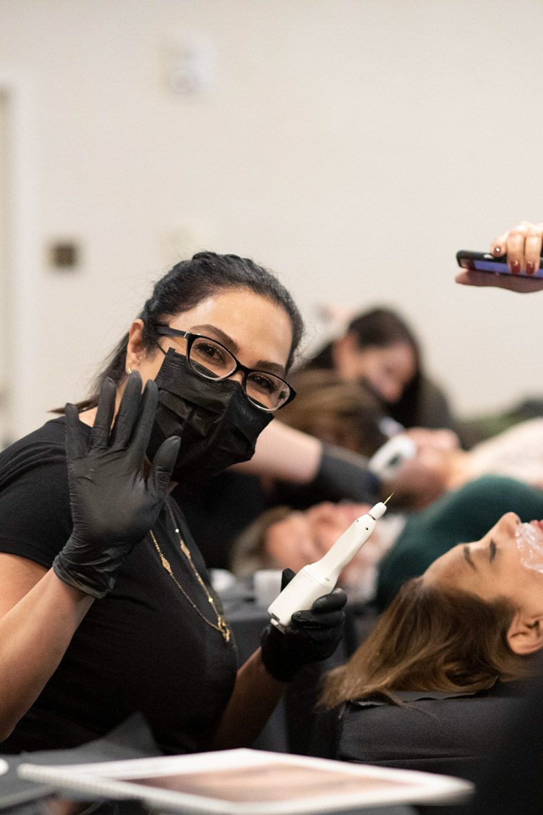 A practitioner holding a plasma pen during a Privé live training, with models on treatment beds behind her